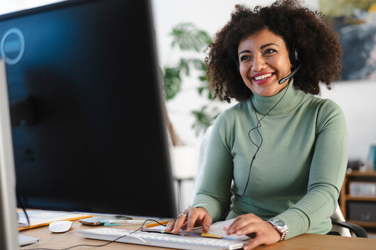 A smiling woman wearing a headset works at her computer, typing on the keyboard and looking at the screen.