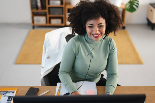 A smiling woman with curly hair wears a headset and works at her desk, looking at a computer screen. She is taking notes in a notebook.