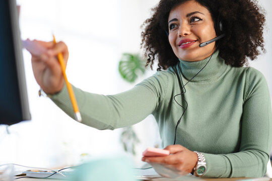 A woman wearing a headset and a green turtleneck smiles as she holds a pencil and a notepad, working at her desk.