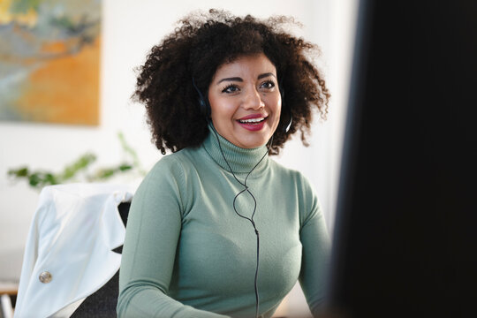 A smiling woman with curly hair wears a headset and a green turtleneck while working at her desk.