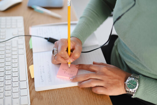 A person is writing notes on a pink sticky note placed on a notebook, with a pencil in hand, at a desk with a keyboard and mouse.