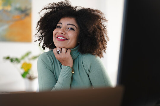 A smiling woman with curly hair wearing a green turtleneck and a large ring on her finger, resting her chin on her hand.