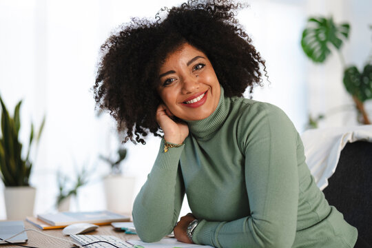 A smiling woman with curly hair rests her head on her hand while sitting at a desk with a computer and plants.