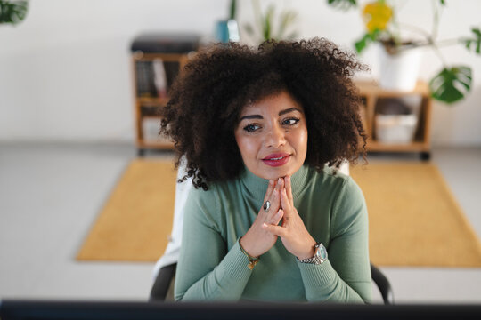 A woman with curly hair sits at a desk, looking thoughtfully into the distance. She rests her chin on her hands, wearing a green turtleneck and jewelry.