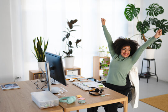 A woman with an afro smiles and raises her arms in triumph while working at her desk in a bright, plant-filled home office.