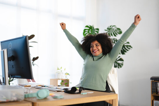 A woman celebrates success at her desk, raising her arms in triumph. She is smiling and looking away from the computer screen.