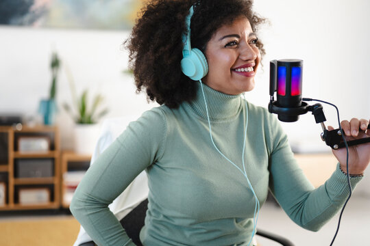 A woman with curly hair wears headphones and speaks into a microphone, recording audio for a podcast or broadcast.