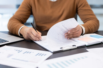 Hands writing with pen on official paperwork beside a laptop while reviewing documents, symbolizing contract signing, administrative work, legal and financial processing in an office setting