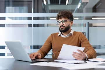 Young businessman analyzing data and typing on a laptop, managing paperwork and responsibilities while staying productive in a modern office environment