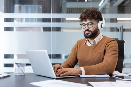 Young indian man in a brown sweater and glasses sitting at a desk, typing on a laptop and wearing white headphones, concentrating on his work in a contemporary office environment