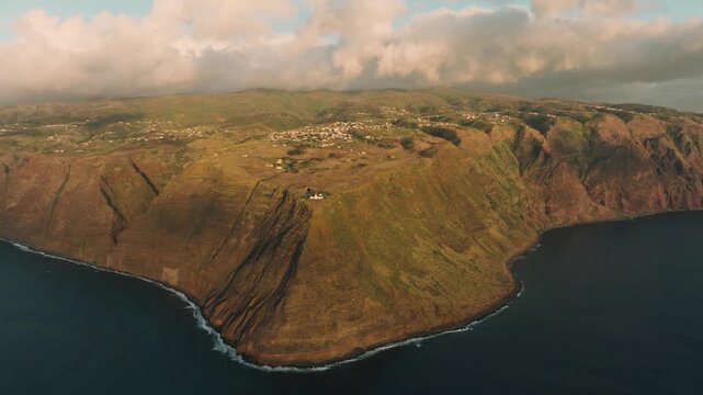 A cinematic drone shot of Farol da Ponta do Pargo, the westernmost lighthouse of Madeira, during sunset. The lighthouse stands prominently on rugged coastal cliffs, with the ocean and dramatic sky 