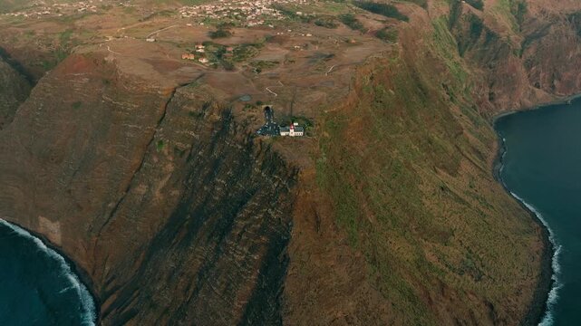 A cinematic drone shot of Farol da Ponta do Pargo, the westernmost lighthouse of Madeira, during sunset. The lighthouse stands prominently on rugged coastal cliffs, with the ocean and dramatic sky 