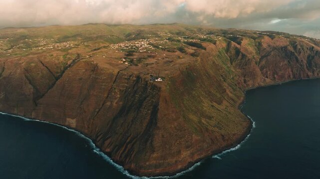 A cinematic drone shot of Farol da Ponta do Pargo, the westernmost lighthouse of Madeira, during sunset. The lighthouse stands prominently on rugged coastal cliffs, with the ocean and dramatic sky 