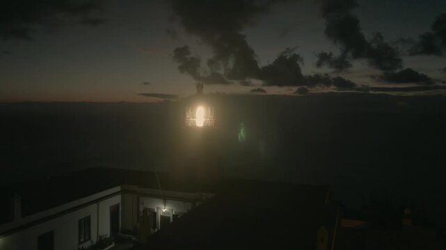 Drone footage of Farol da Ponta do Pargo lighthouse on Madeira Island at night. The red and white lighthouse rotates its powerful beacon across the dark ocean, with beams of light sweeping directly in