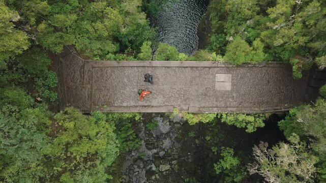 A static top-down drone shot of two male hikers walking across a historic stone bridge over a flowing river on the Levada das 25 Fontes trail in Madeira. Heavy rain falls, creating a dramatic
