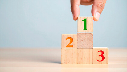 Hand placing a wooden block labeled &ldquo;1&rdquo; atop two stacked blocks labeled &ldquo;2&rdquo; and &ldquo;3&rdquo; featuring a podium. The image symbolizes prioritization, competitiveness, achieving budgets in the business world.