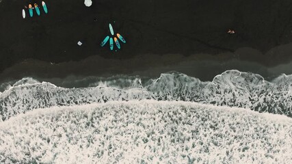 A smooth vertical aerial drone descent over a black volcanic beach in Seixal, Madeira. The camera looks straight down, revealing dark volcanic sand shaped by ancient lava flows, with ocean waves 