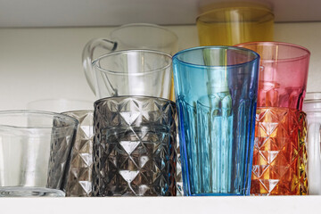 Colorful drinking glasses on kitchen shelf. Transparent blue orange and pink glassware collection. Modern home interior detail. Clean empty cups arranged in row. Household design background.