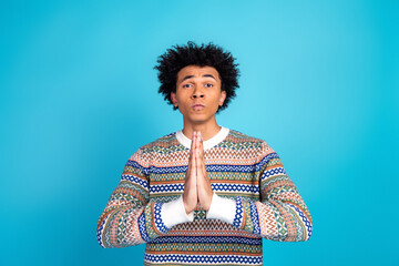 Young man in colorful sweater stands against blue background with hands pressed together in prayer gesture for stock photography and lifestyle advertising