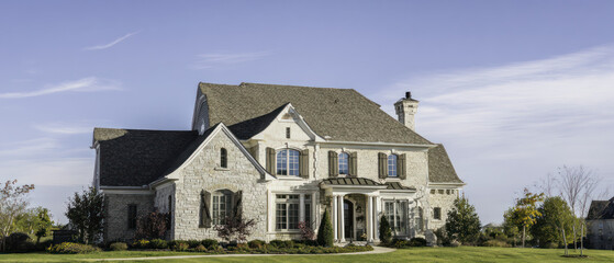 Spacious country house with pale stone walls, arched shutters, manicured garden, and classical columns under a soft sky.
