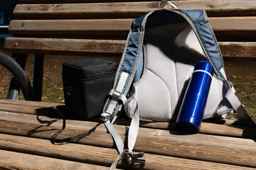 A blue and gray backpack, a black square case, and a blue metallic thermos are resting on a slatted...