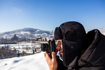 Profile portrait of a winter photographer holding a camera to his eye, black hooded coat, snow covered hills and cityscape behind, clear sky, cold travel scene outdoors. © Ernad