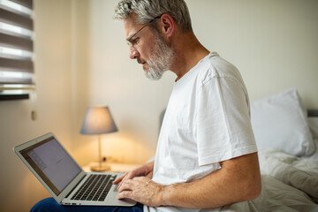 Mature man focused on laptop while working in bedroom