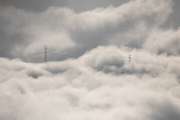 Winter mountain dawn, where a high-voltage pylon rises through rolling clouds and fog, silhouetted against soft morning light.