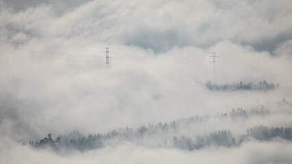 Winter mountain dawn, where a high-voltage pylon rises through rolling clouds and fog, silhouetted against soft morning light.
