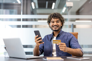 Portrait of a young Indian man sitting at his workplace in the office, holding a credit card and phone, looking at the camera