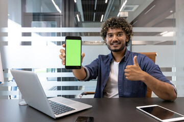 Portrait of an Indian young man in an office, sitting at a desk and showing a green screen mockup of a mobile phone to the camera