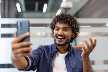 Close-up photo of a young Indian man indoors talking on the camera of a mobile phone he is holding