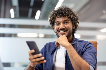Fototapeta na wymiar Close-up portrait of a young Indian man sitting in a chair at a desk in an office, holding a phone and smiling at the camera
