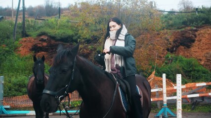 A happy young caucasian woman with dark hair sits astride a docile stallion and puts on gloves in a rural landscape on an autumn day. Horseback riding training for active recreation enthusiasts.
