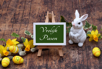 De tekst Vrolijk Pasen staat geschreven op een schildersezel met paasversieringen en bloemen.