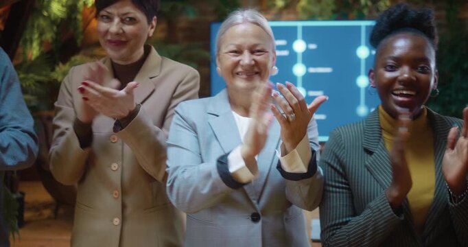 Multicultural business team standing and applauding while looking at camera, smiling as camera pans left to right, capturing each person celebrating success in modern eco-friendly conference room.