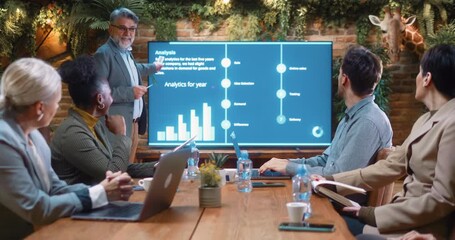 Senior business manager presenting company analytics and performance data on TV screen during corporate meeting, explaining strategy and results to colleagues in modern office. - Powered by Adobe