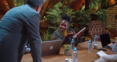 African businesswoman using tablet and communicating with team leader during corporate meeting, discussing data and ideas in modern eco conference room with greenery. - Powered by Adobe