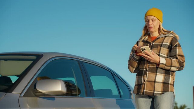 Annoyed young woman standing next to broken down car on side of road, anxiously calling for help and roadside assistance service while gesturing in frustration