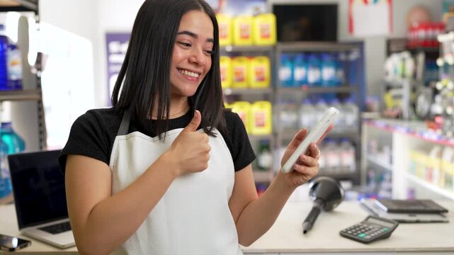 Cheerful young latin woman wearing an apron joyfully gesturing while using a smartphone in a store
