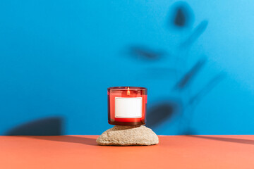 Scented red candle in glass jar placed on stone against blue and coral background. Minimal decor style, mockup