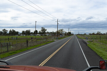 Fototapeta premium A long rural road stretches out in front of me, bordered by expansive fields and tall power lines looming overhead, all beneath a moody overcast sky