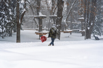 A worker walking through deep snow carrying a red shovel near an aerial rope park structure and bare trees