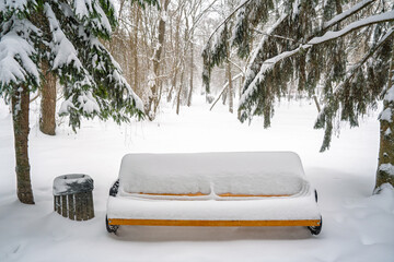 A wooden park bench completely covered by a thick layer of fresh white snow in a cold forest landscape.