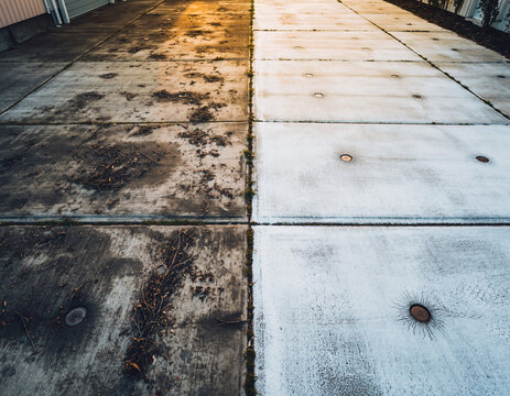 The dirty garage driveway is divided into two parts showcasing the transformation from a stained concrete pathway to a clean, bright, and pristine white surface following a pressure washing service. - Powered by Adobe