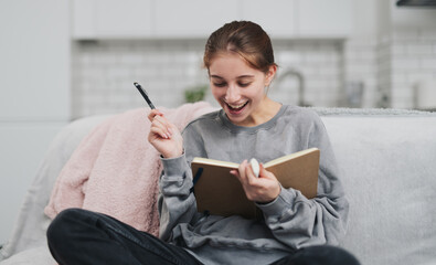 Happy, Smiling, Pretty Girl Writing Funny Notes In A Notebook While Sitting On A Sofa At Home