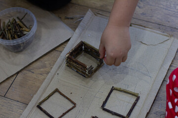 A child's hand builds a small imaginary house using natural wooden sticks and glue on a cardboard...