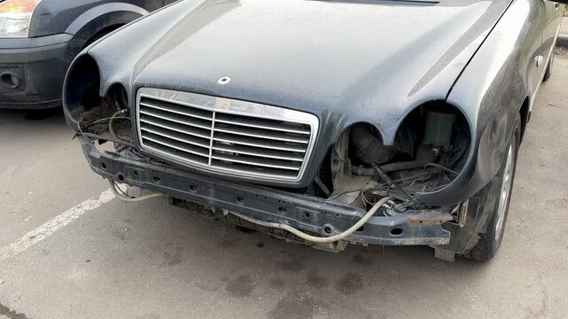 Damaged Car Front End Exposed Bumper Parked On Cracked Asphalt In Urban Lot, Missing Headlights, Twisted Metal, Scratched Paint, Exposed Wiring, Bent Grille, Adjacent Sedan Visible.