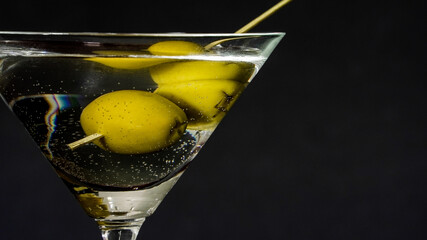 Clear glass containing cocktail with olives on a metal skewer against dark background