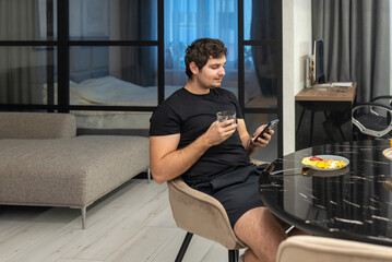 Young man checking smartphone while holding glass of water at breakfast